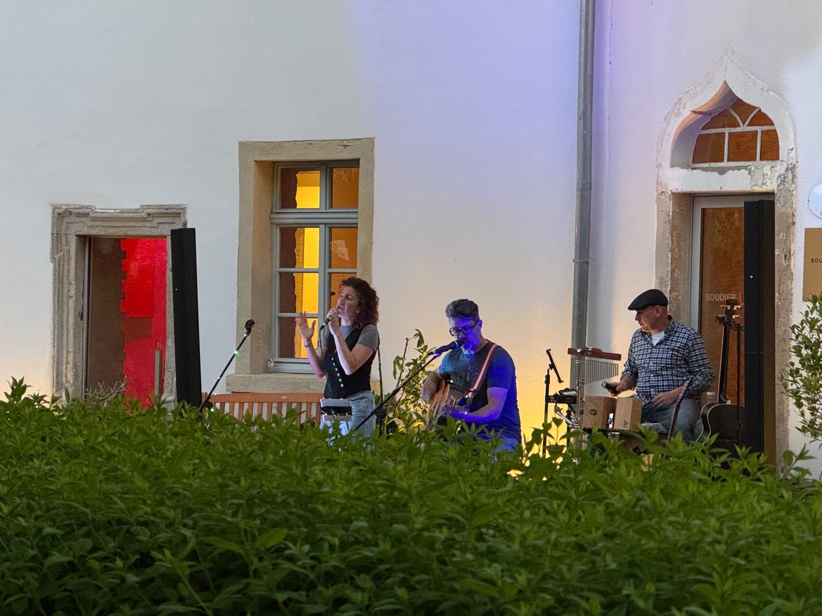 Drei Personen bei einem Live-Konzert im Freien vor einer weißen Hauswand mit drei Fenstern, eine Frau singt, ein Mann spielt Gitarre und ein weiterer Mann spielt Cajón, im Vordergrund grüne Pflanzen