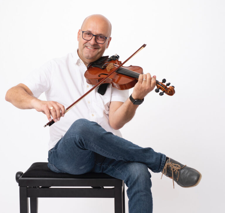 Mann mit Brille und weißem Hemd sitzt mit verschränkten Beinen auf einem schwarzen Klavierhocker und spielt Geige vor weißem Hintergrund