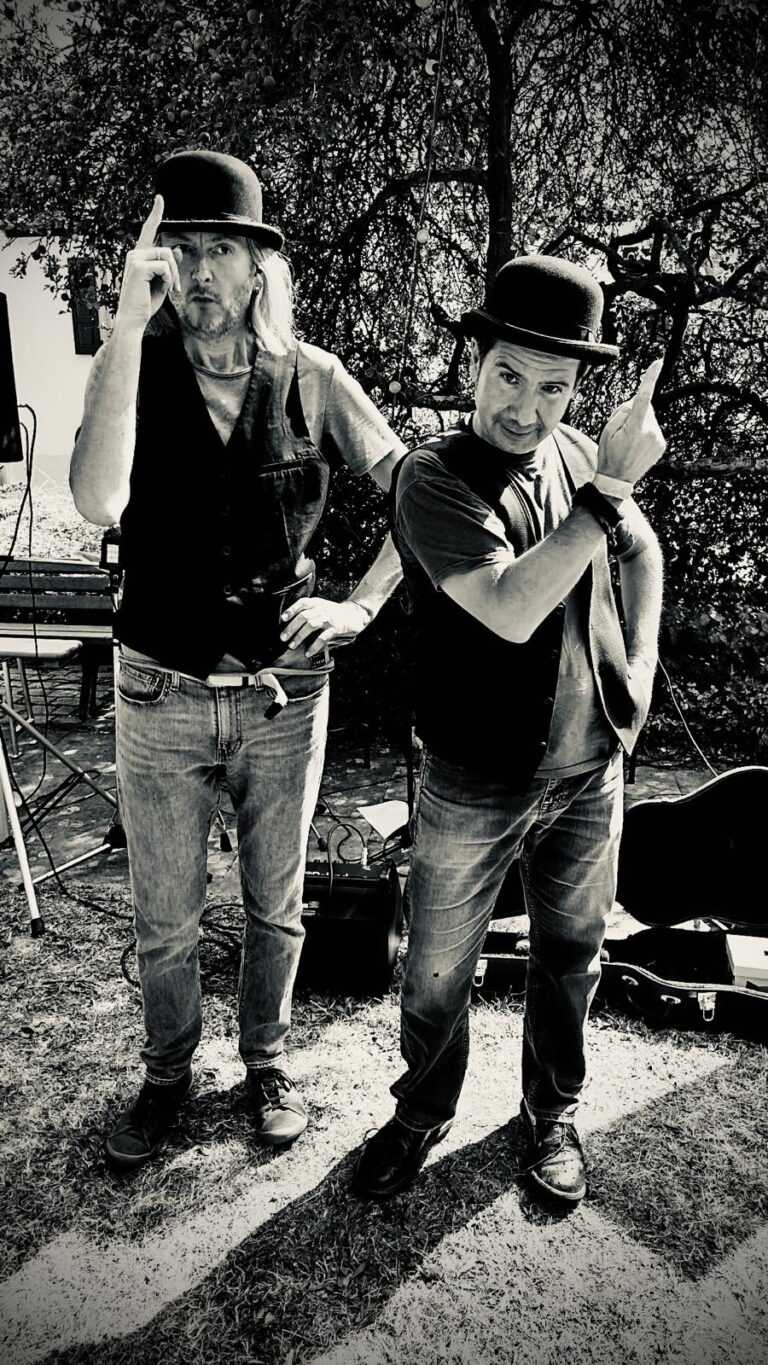 Schwarzweiß-Foto von zwei Männern mit schwarzen Melonenhüten, Jeans und Westen, die vor einem Baum stehen und jeweils mit einem Finger nach oben zeigen, im Hintergrund Gartenmöbel und Musikinstrumentenkoffer.