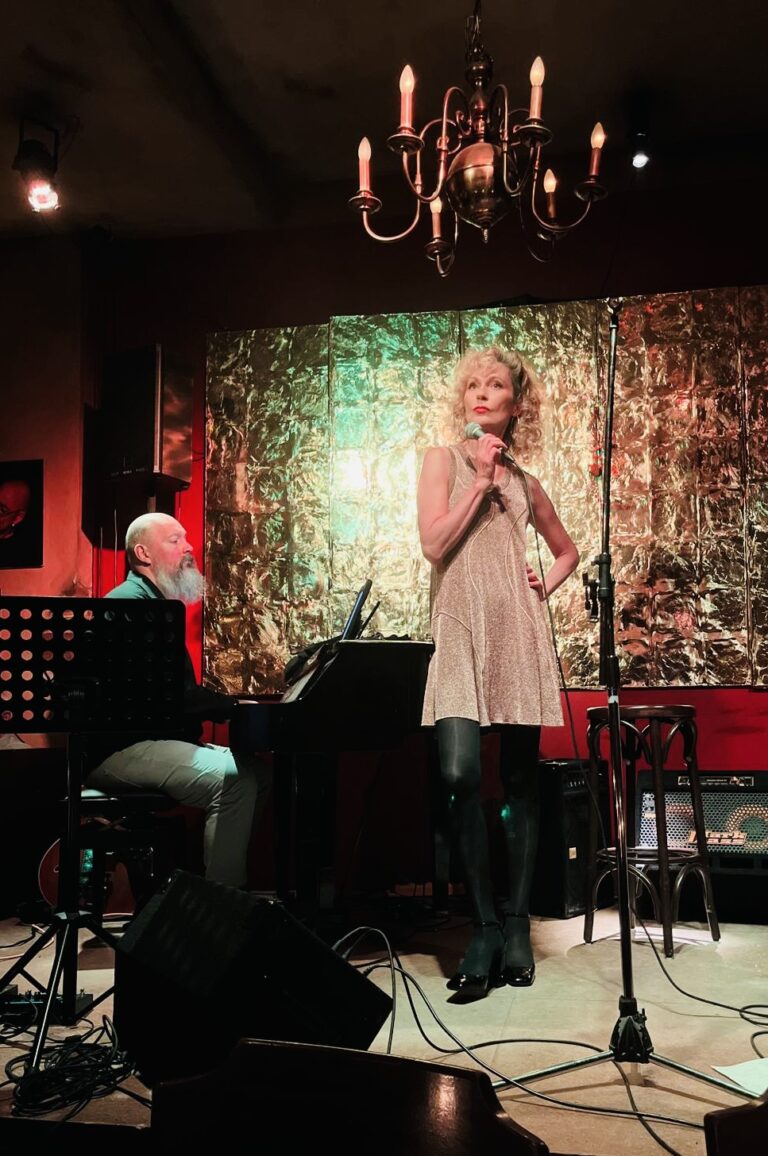 Eine Frau mit lockigem, blonden Haar und einer silbernen Kleid steht auf einer Bühne und hält ein Mikrofon, während ein Mann mit bart und Glatze am Flügel Klavier spielt, im Hintergrund eine strukturierte, metallic wirkende Wand und ein Kronleuchter an der Decke.