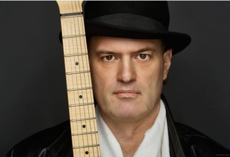 Porträt eines Mannes mit Hut und Lederjacke, der ernst in die Kamera schaut, mit dem Hals eines Gitarrenhalses, der sein Gesicht teilweise verdeckt.