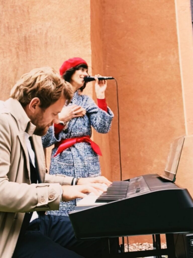 Ein Mann spielt Klavier, während eine Frau mit roter Mütze und blauem Mantel daneben singt und ein Mikrofon hält, vor einer orangefarbenen Wand.