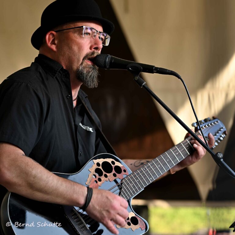 Mann mit schwarzem Hut, Brille und Bart singt in ein Mikrofon und spielt eine Akustikgitarre mit auffälligen Schalllöchern auf einer Bühne.