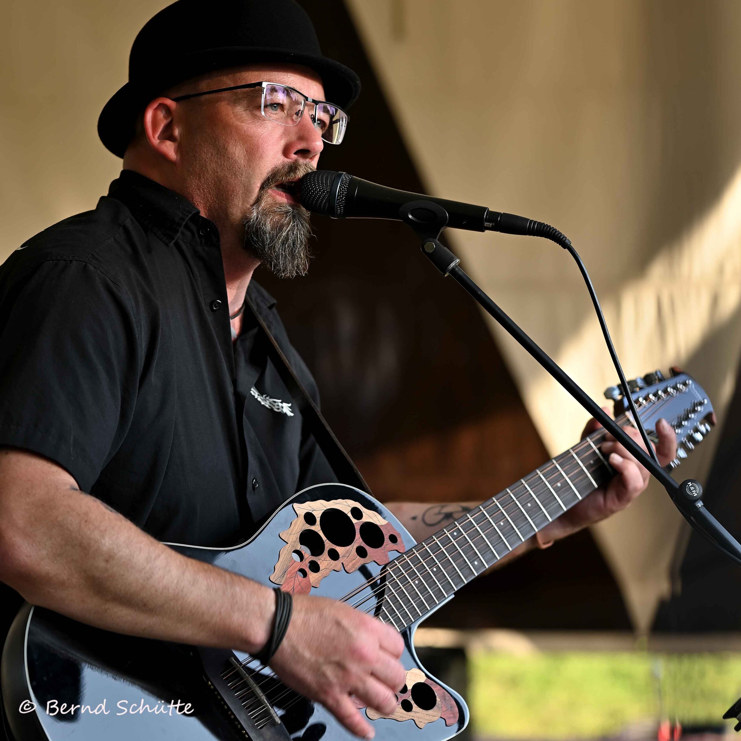 Mann mit schwarzem Hut, Brille und Bart singt in ein Mikrofon und spielt eine Akustikgitarre mit auffälligen Schalllöchern auf einer Bühne.