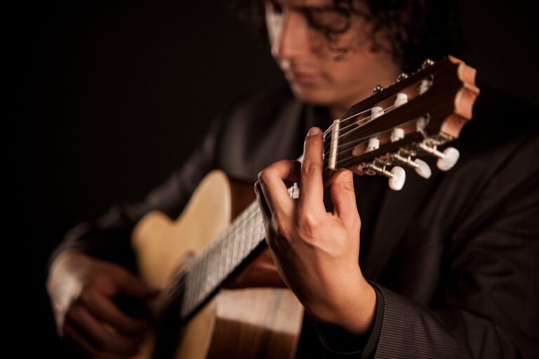 Mann mit dunklem Anzug, der eine klassische Gitarre spielt, Nahaufnahme seiner linken Hand, die die Saiten auf dem Gitarrenhals drückt, vor dunklem Hintergrund