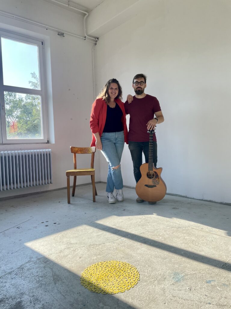 Ein Mann und eine Frau stehen in einem hellen, leeren Raum mit Betonboden; die Frau trägt eine rote Jacke und eine blaue Jeans, der Mann hält eine akustische Gitarre und hat eine Brille, neben ihnen steht ein leerer Holzstuhl, Sonnenlicht fällt durch ein Fenster auf den Boden mit einem gelben dekorativen Objekt.