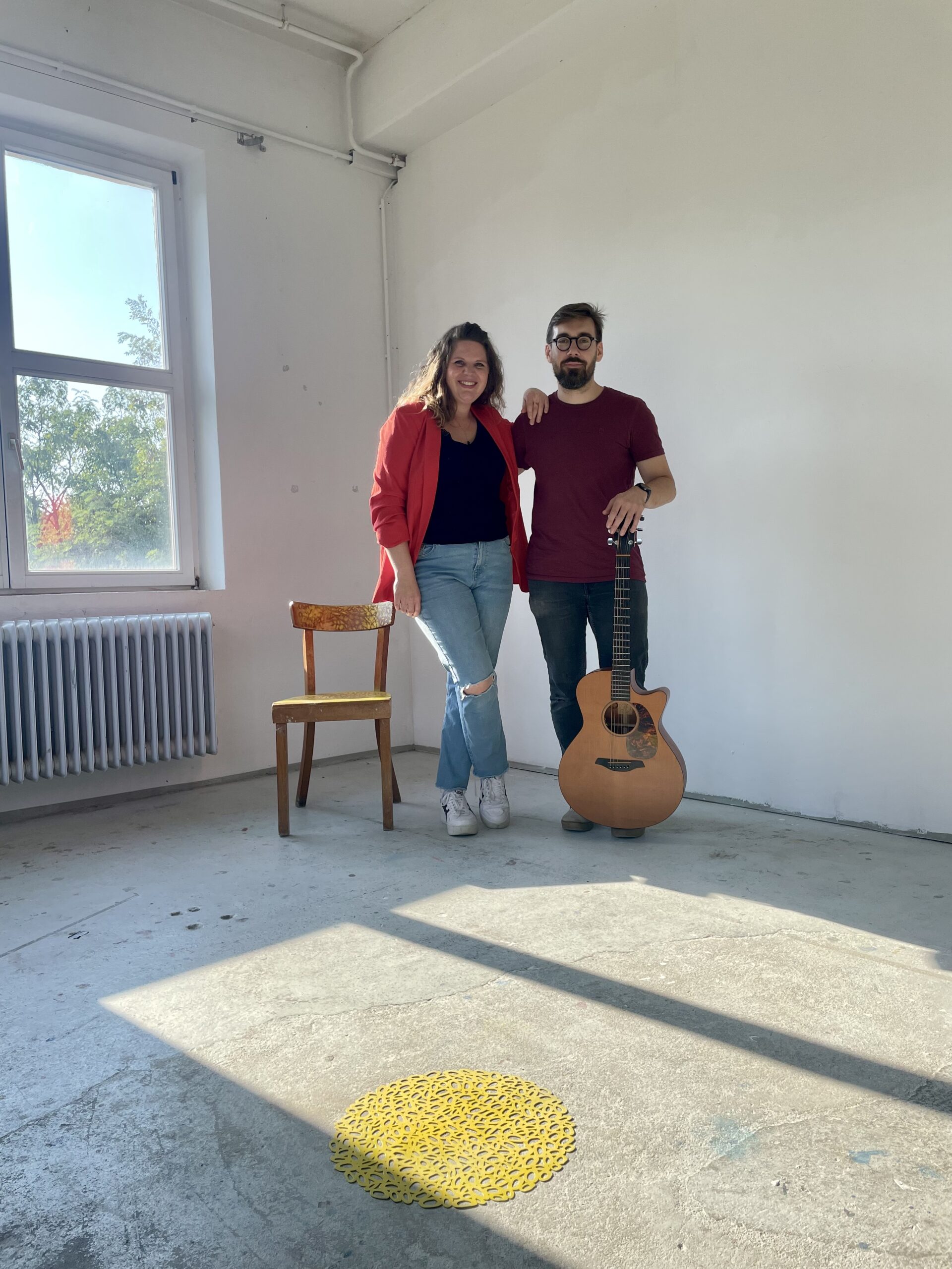 Ein Mann und eine Frau stehen in einem hellen, leeren Raum mit Betonboden; die Frau trägt eine rote Jacke und eine blaue Jeans, der Mann hält eine akustische Gitarre und hat eine Brille, neben ihnen steht ein leerer Holzstuhl, Sonnenlicht fällt durch ein Fenster auf den Boden mit einem gelben dekorativen Objekt.