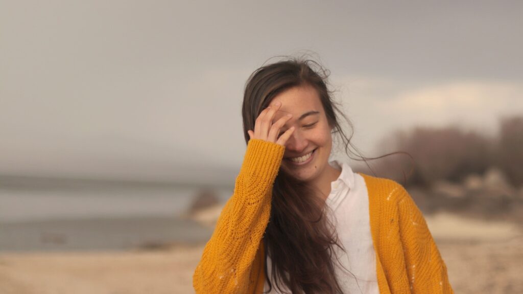 Su-Min: Junge Frau mit langem braunem Haar und gelbem Strickcardigan, die am Strand steht und mit geschlossenen Augen lächelt, während sie eine Hand an die Stirn hält.