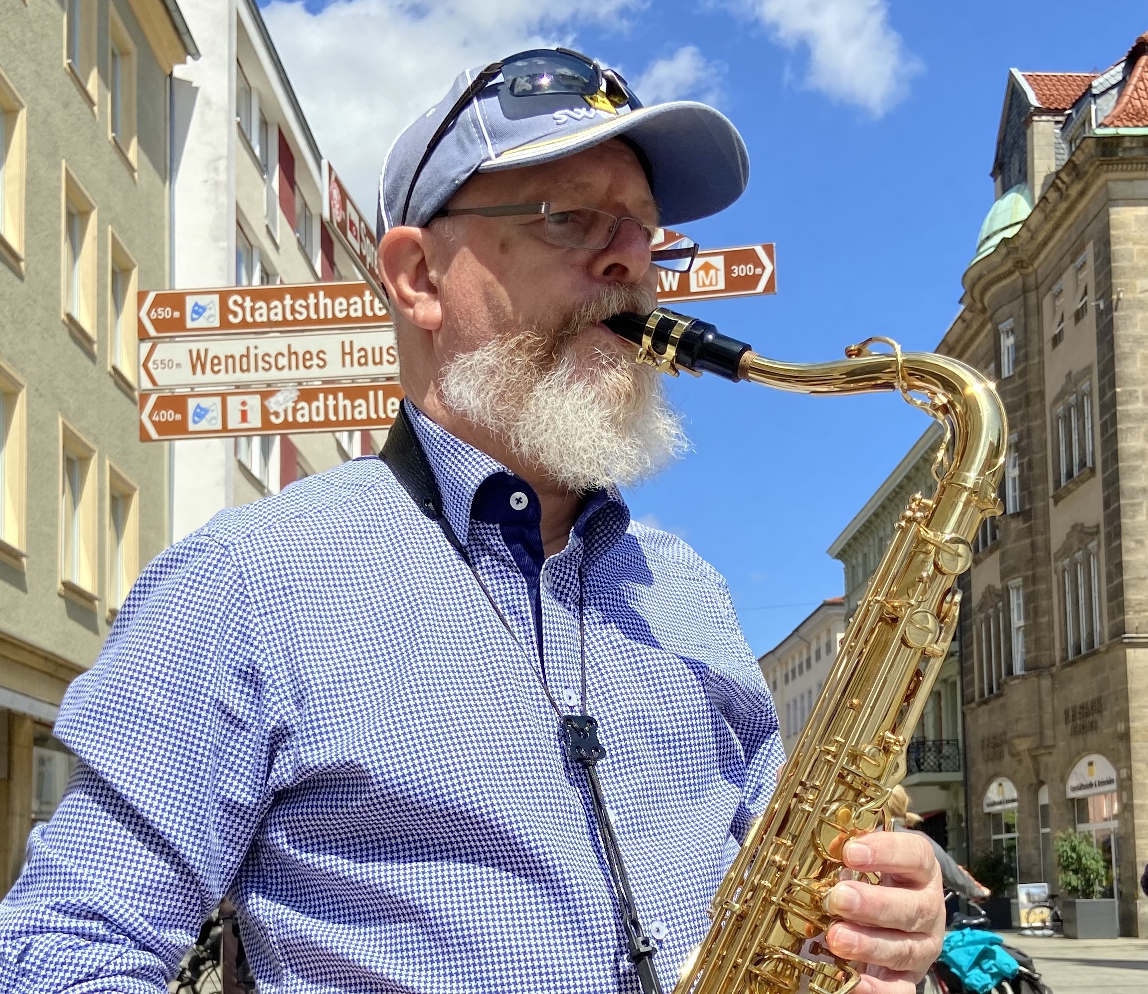 Älterer Mann mit weißem Bart und blauer Kappe spielt Saxophon in einer sonnigen Stadtstraße, im Hintergrund Wegweiser zu Staatsheater, Wendisches Haus und Stadthalle.