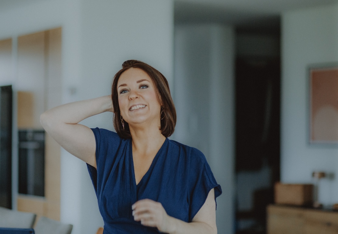 Monika Erdmann: Frau mit kurzen braunen Haaren und blauem Oberteil, die lächelnd eine Hand hinter den Kopf legt, in einem modernen Innenraum mit unscharfem Hintergrund.
