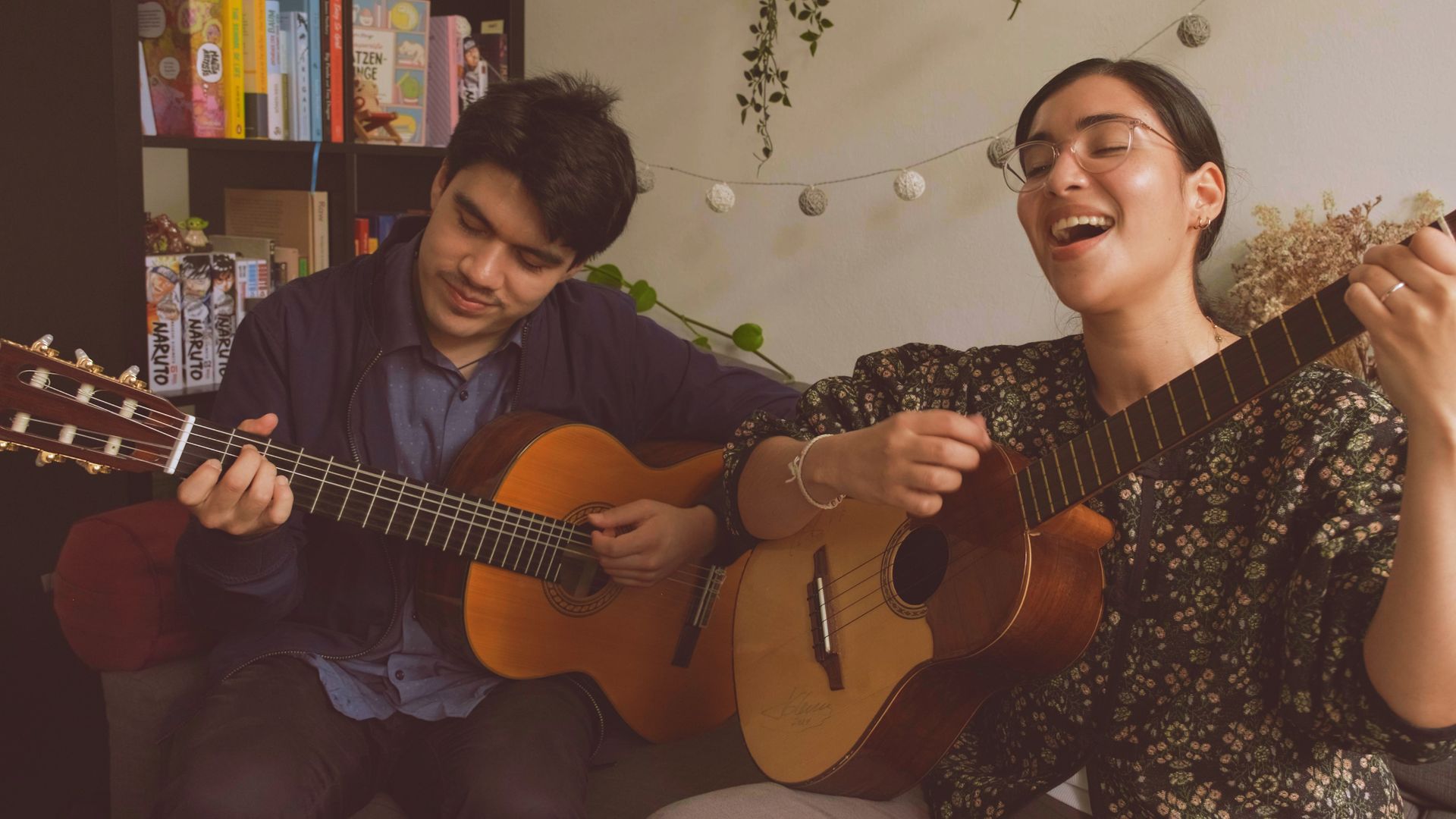 Ein Mann und eine Frau sitzen auf einem Sofa, spielen Gitarren, die Frau singt und hat eine Brille und ein geblümtes Oberteil, im Hintergrund ein Bücherregal und eine Lichterkette an der Wand.