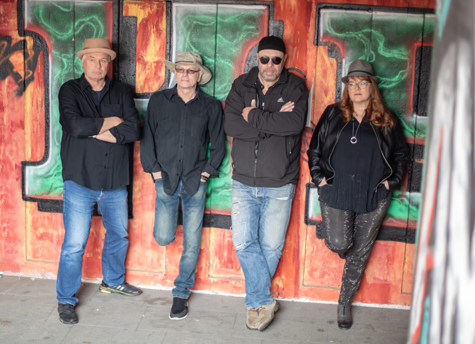 FreiHerr: Vier Personen lehnen vor einer orange-grün-schwarzen Graffiti-Wand, alle tragen Hüte, drei Männer in Freizeitkleidung und eine Frau in schwarzer Jacke und Glitzerhose, alle mit ernstem Gesichtsausdruck.