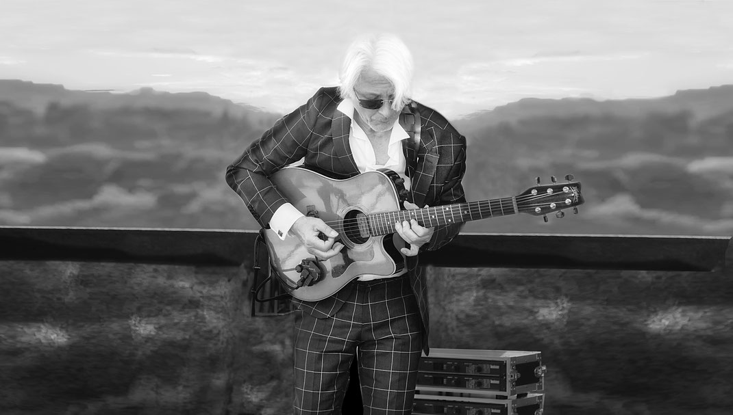 Peter Hillert : Schwarz-weiß-Foto eines älteren Mannes mit weißem Haar und Sonnenbrille, der im Anzug mit Karomuster eine Akustikgitarre spielt vor verschwommenem Hintergrund mit Equipment-Koffern.