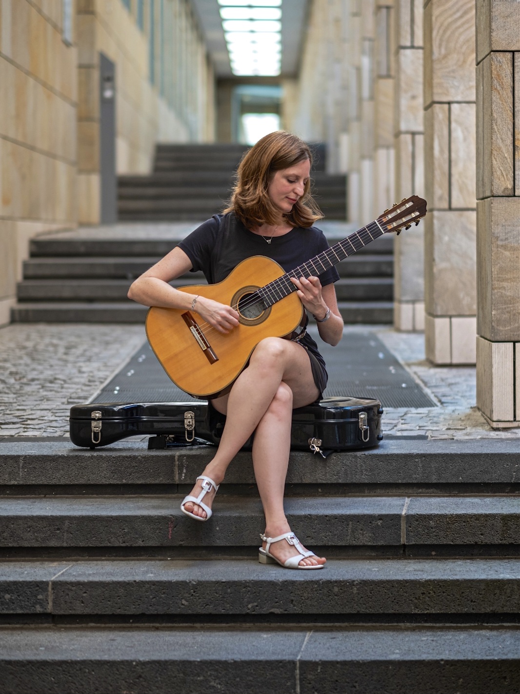 Frau mit braunen Haaren in schwarzem T-Shirt und schwarzem Rock, die auf einer Treppenstufe sitzt und klassische Gitarre spielt, im Hinterhof mit Sandsteinmauern und Treppen im Hintergrund.