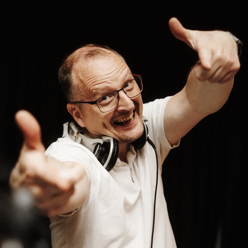 DJ Chaques: Mann mit Brille und Kopfhörern um den Hals zeigt lachend zwei Daumen nach oben vor schwarzem Hintergrund