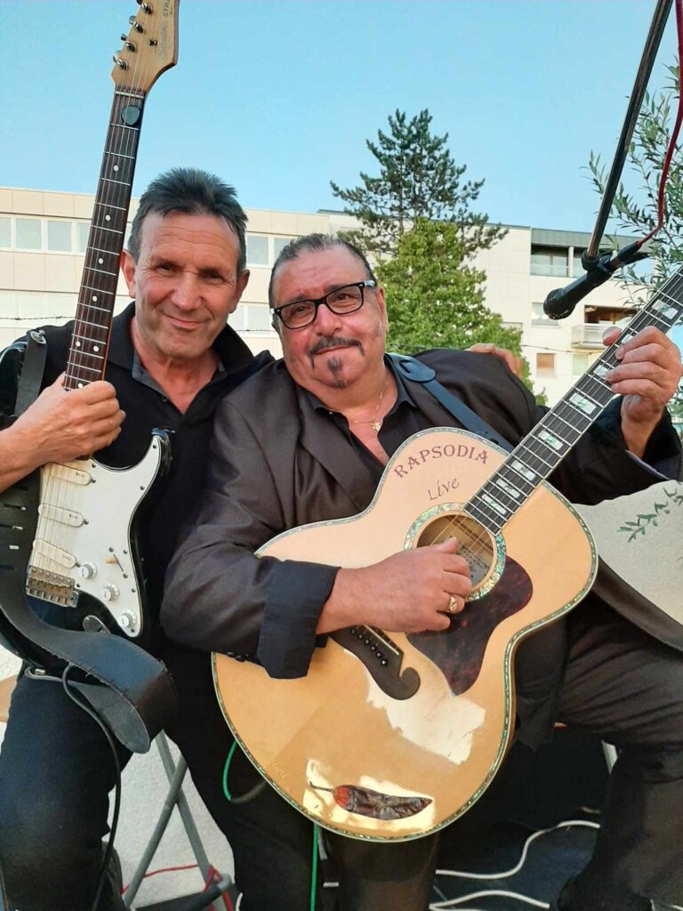 Rapsodia-live: Zwei Männer sitzen nebeneinander und halten Gitarren, der linke Mann hält eine elektrische Gitarre, der rechte eine akustische Gitarre mit der Aufschrift "RAPSODIA Live", im Hintergrund ein Baum und ein Gebäude bei Tageslicht.