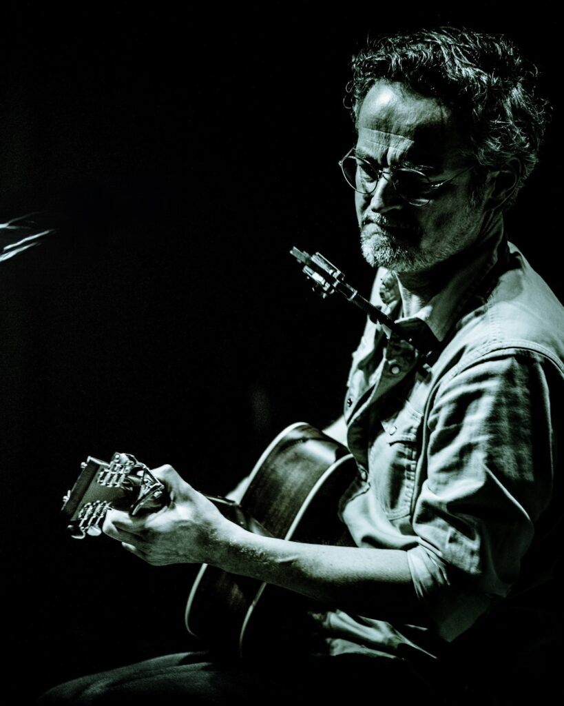 Maik Garthe: Mann mit Brille und leichtem Bart spielt Gitarre, in gedämpftem Licht vor dunklem Hintergrund, konzentrierter Gesichtsausdruck.