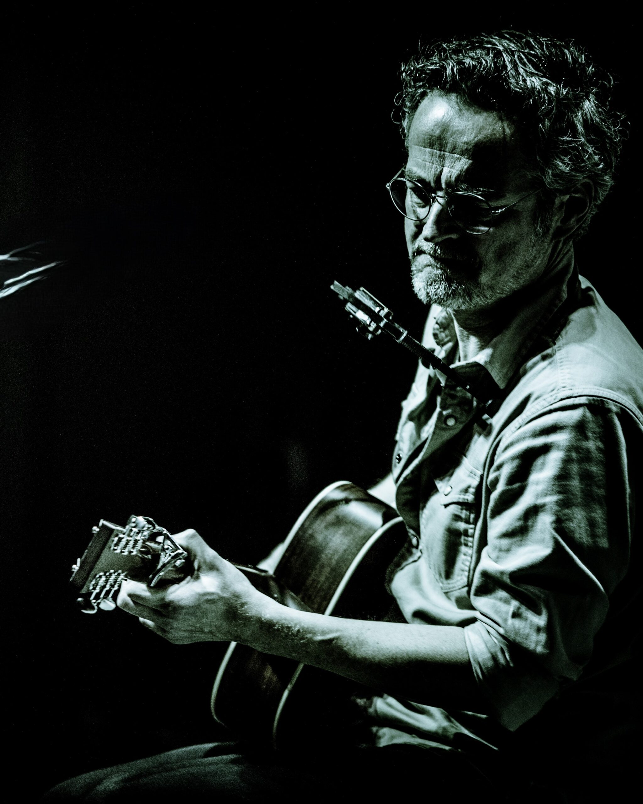 Maik Garthe: Mann mit Brille und leichtem Bart spielt Gitarre, in gedämpftem Licht vor dunklem Hintergrund, konzentrierter Gesichtsausdruck.