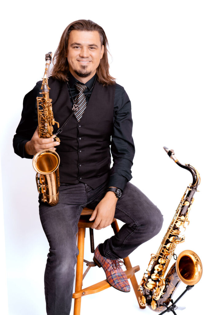 Mann mit langen Haaren und grüner Krawatte sitzt auf Hocker und hält ein goldenes Altsaxophon, daneben steht ein Baritonsaxophon.