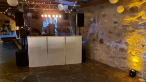 Dunkler Raum mit DJ-Pult, Lautsprechern, dekorativen Lichterketten und gelben Lampions an einer rustikalen Wand aus Stein.