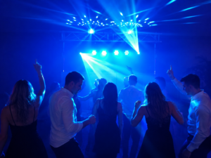 Gruppe von Menschen in eleganter Kleidung tanzt vor einer Bühne mit blauem Bühnenlicht und Nebeleffekt.