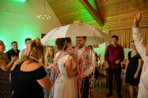 Braut und Bräutigam unter einem großen weißen Regenschirm umgeben von Gästen, alle mit bunten Partybändern geschmückt in einem festlich beleuchteten Raum.