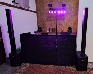 DJ-Pult mit Lautsprecherboxen und violetter Bühnenbeleuchtung in einem Raum mit Teppichboden und Holzregal an der Wand