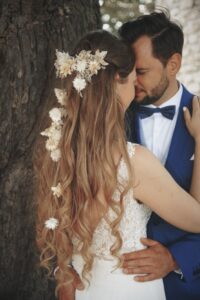 Nahaufnahme eines Brautpaars in inniger Umarmung vor einem Baum, die Braut trägt ein weißes Spitzenkleid mit langen lockigen Haaren, die mit weißen getrockneten Blumen geschmückt sind, der Bräutigam trägt einen blauen Anzug mit weißem Hemd und schwarzer Fliege.