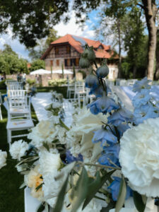 Nahaufnahme eines Blumenarrangements mit weißen und blauen Blüten vor einer Außenkulisse mit weißen Stühlen und einem Fachwerkhaus mit rotem Dach unter blauem Himmel.