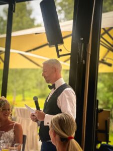 Mann in weißem Hemd, schwarzer Weste und Fliege hält ein Mikrofon und Papier, während er spricht, links davon sitzt eine lächelnde Frau in einem ärmellosen Spitzenkleid, im Hintergrund gelbe Sonnenschirme und grünes Außenambiente.