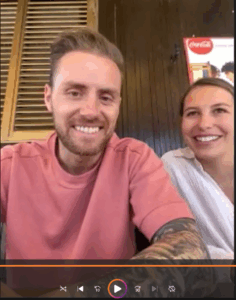Zwei lächelnde Personen, ein Mann mit rosa Oberteil und tätowiertem Arm links, eine Frau in weißem Hemd rechts, vor einer dunklen Holzwand mit einem Coca-Cola-Poster