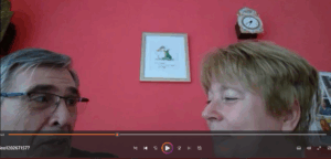 Nahaufnahme eines Mannes mit grauem Haar und Brille, der eine Frau mit kurzem blonden Haar ansieht, vor einer roten Wand mit einer gerahmten Zeichnung und einer Wanduhr im Hintergrund.