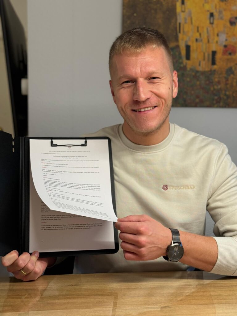 Ein lächelnder Mann mit kurzem blonden Haar trägt einen hellen Pullover und hält einen Klemmbrett-Ordner mit mehreren beschriebenen Blättern an einem Holztisch, im Hintergrund ist ein Gemälde an der Wand zu sehen.