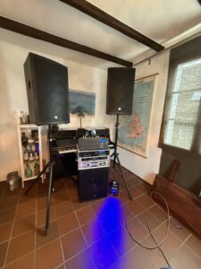 Musikanlage mit zwei Lautsprechern auf Stativen, Verstärker, und Mischpult vor einem schwarzen Klavier in einem Raum mit braunem Fliesenboden, weißen Wänden, einer Landkarte der Britischen Inseln und einem beleuchteten Schrank mit Flaschen.