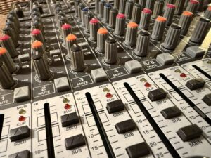 Nahaufnahme eines Mischpults mit mehreren Reglern, Schiebeschaltern und Drehknöpfen in verschiedenen Farben zur Lautstärke- und Klangregelung.