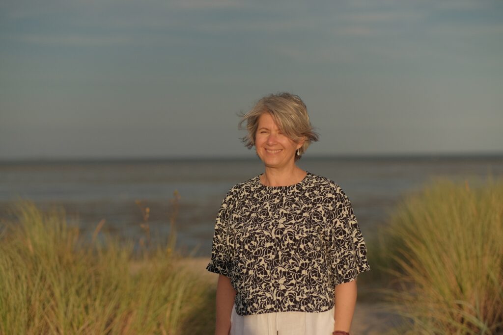 Ältere Frau mit kurzen grauen Haaren und gemustertem Oberteil, die lächelnd im Freien zwischen Strandgräsern steht, mit einem Gewässer und wolkigem Himmel im Hintergrund.