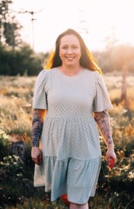 Frau mit langen braunen Haaren, die ein hellblaues, kurzärmeliges Kleid mit Lochmuster trägt und Tattoos an beiden Unterarmen zeigt, steht lächelnd in einer sonnenbeschienenen Wiesenlandschaft am späten Nachmittag.